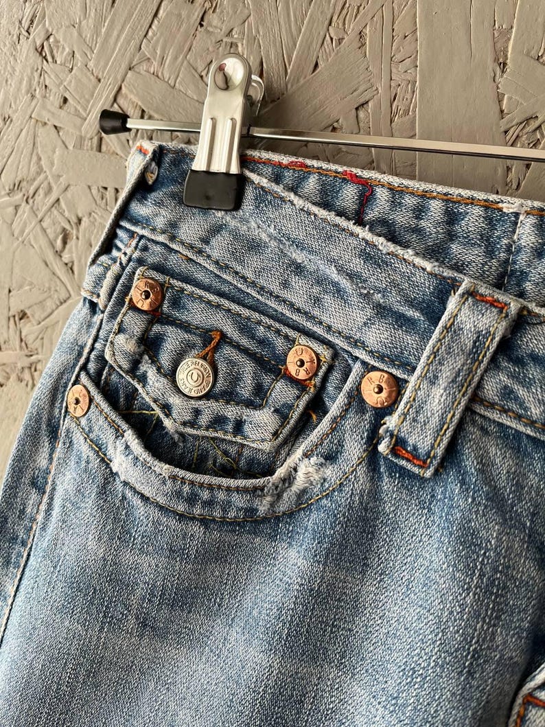 Puede incluir: Primer plano de unos vaqueros de mezclilla azul claro con remaches de color cobre y un peque&ntilde;o bolsillo delantero. Los vaqueros est&aacute;n desgastados, mostrando signos de uso. Las costuras son de color naranja en contraste. Los vaqueros cuelgan de una percha met&aacute;lica.