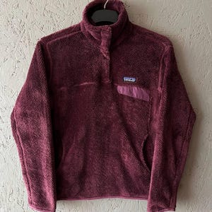 May include: A burgundy fleece pullover with a high collar and a small chest pocket. The pullover has a button closure at the neck and a small brand patch on the chest pocket. The fleece material appears soft and textured.