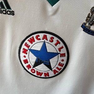 K&ouml;nnte beinhalten: Nahaufnahme eines wei&szlig;en Fu&szlig;balltrikots mit gr&uuml;nen Akzenten und dem Adidas-Logo. Das Trikot zeigt einen runden Aufn&auml;her mit der Aufschrift "NEWCASTLE BROWN ALE" um einen blauen Stern und eine Silhouette.