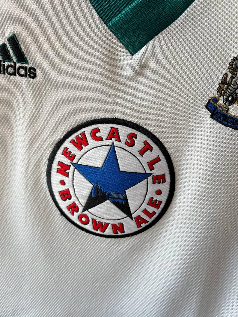 K&ouml;nnte beinhalten: Nahaufnahme eines wei&szlig;en Fu&szlig;balltrikots mit gr&uuml;nen Akzenten und dem Adidas-Logo. Das Trikot zeigt einen runden Aufn&auml;her mit der Aufschrift "NEWCASTLE BROWN ALE" um einen blauen Stern und eine Silhouette.