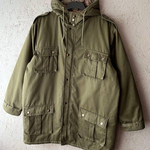 K&ouml;nnte beinhalten: Eine olivgr&uuml;ne Feldjacke mit Kapuze und Kordelzug, einem Frontrei&szlig;verschluss mit Druckknopfleiste, zwei Brusttaschen mit Klappen sowie zwei unteren Taschen mit Klappen. Die Jacke ist mit Schulterklappen und langen &Auml;rmeln ausgestattet.