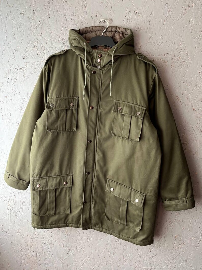 K&ouml;nnte beinhalten: Eine olivgr&uuml;ne Feldjacke mit Kapuze und Kordelzug, einem Frontrei&szlig;verschluss mit Druckknopfleiste, zwei Brusttaschen mit Klappen sowie zwei unteren Taschen mit Klappen. Die Jacke ist mit Schulterklappen und langen &Auml;rmeln ausgestattet.