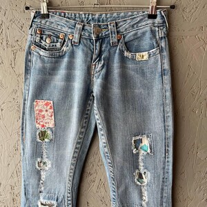 Puede incluir: Vaqueros de mezclilla azul claro con parches decorativos y detalles desgastados. Los vaqueros cuelgan de una percha de metal. Incluyen cierre de bot&oacute;n, bolsillos y un lavado descolorido.
