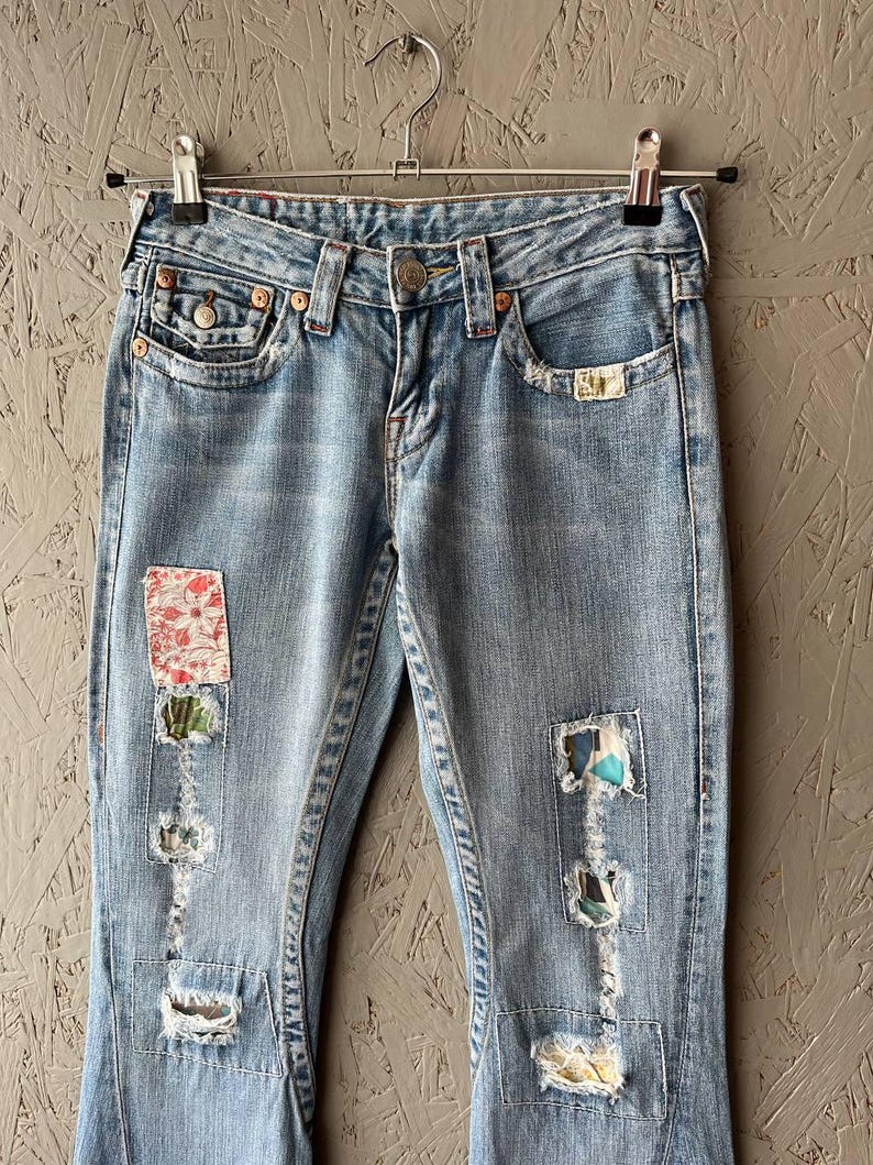 Puede incluir: Vaqueros de mezclilla azul claro con parches decorativos y detalles desgastados. Los vaqueros cuelgan de una percha de metal. Incluyen cierre de bot&oacute;n, bolsillos y un lavado descolorido.