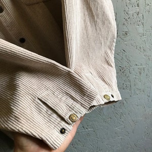 May include: A beige corduroy shirt with a button-up closure and a pocket on the left chest. The shirt has a woven label with the text "Wool" on the left side of the collar.