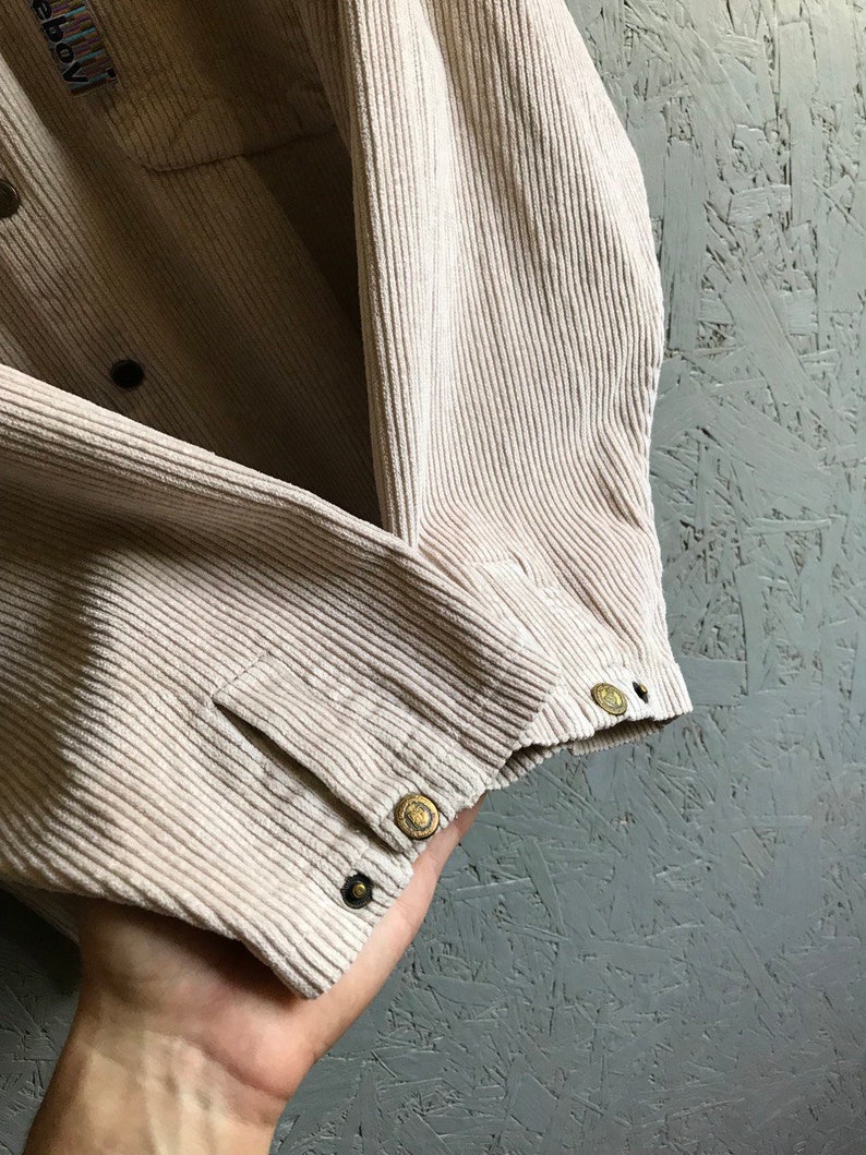 May include: A beige corduroy shirt with a button-up closure and a pocket on the left chest. The shirt has a woven label with the text "Wool" on the left side of the collar.
