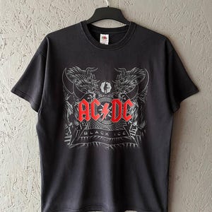 Puede incluir: Camiseta negra con el logo de la banda AC/DC en rojo y blanco. El diseño incluye una ilustración estilizada con las palabras "BLACK ICE" debajo del logo. La camiseta está colgada en una percha negra.