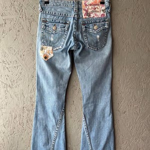 Puede incluir: Vaqueros de mezclilla azul claro con dise&ntilde;o de pernera acampanada. Los vaqueros presentan detalles desgastados, parches decorativos y un parche floral en el bolsillo trasero. La etiqueta de la marca True Religion es visible.