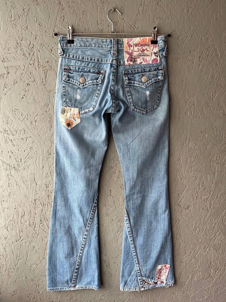 Puede incluir: Vaqueros de mezclilla azul claro con dise&ntilde;o de pernera acampanada. Los vaqueros presentan detalles desgastados, parches decorativos y un parche floral en el bolsillo trasero. La etiqueta de la marca True Religion es visible.