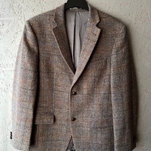 Peut inclure: Un blazer en tweed marron avec un motif à carreaux subtil. La veste présente un revers cranté, une fermeture à deux boutons et une poche poitrine unique. Le blazer est suspendu à un cintre noir sur un fond neutre.