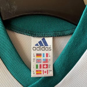 K&ouml;nnte beinhalten: Nahaufnahme eines wei&szlig;en und gr&uuml;nen Adidas-Trikots. Das Trikot hat einen V-Ausschnitt und das Adidas-Logo ist auf einem Etikett mit L&auml;nderflaggen zu sehen. Der Kragen und die Zierleiste sind dunkelgr&uuml;n.