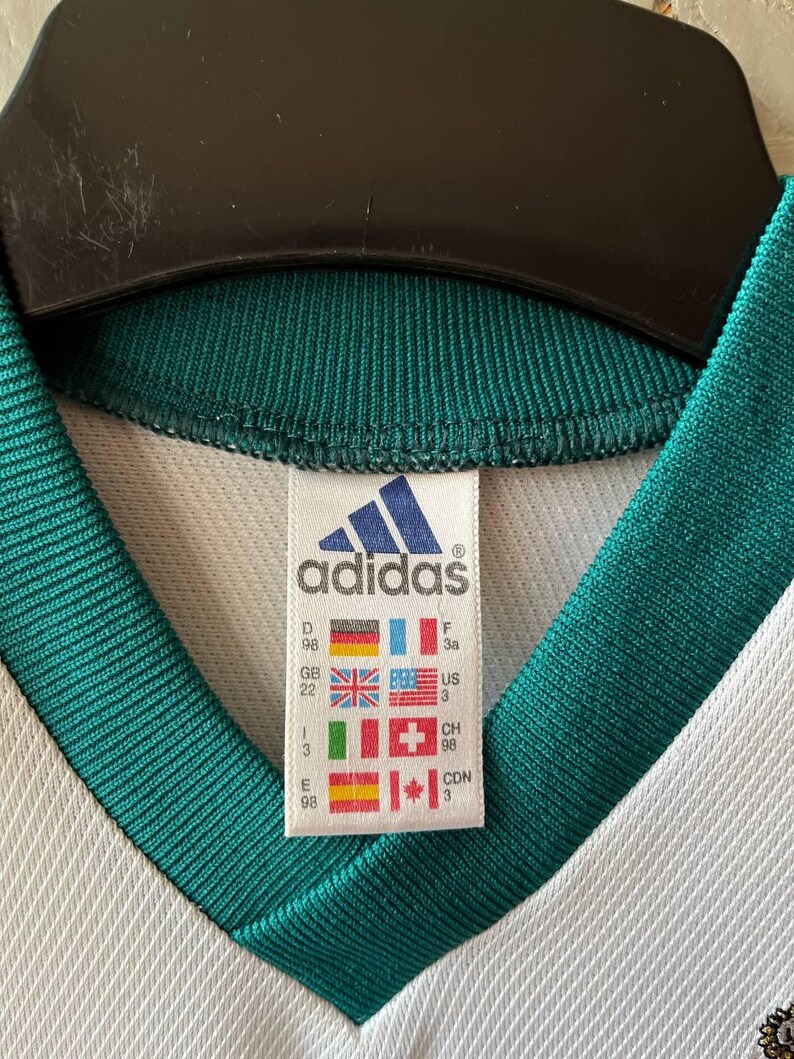 K&ouml;nnte beinhalten: Nahaufnahme eines wei&szlig;en und gr&uuml;nen Adidas-Trikots. Das Trikot hat einen V-Ausschnitt und das Adidas-Logo ist auf einem Etikett mit L&auml;nderflaggen zu sehen. Der Kragen und die Zierleiste sind dunkelgr&uuml;n.