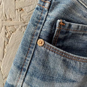 以下が含まれることがあります： 色あせたウォッシュ加工のブルーデニムジーンズのクローズアップ。ジーンズは銅色のステッチと小さなポケットが特徴です。真鍮製のボタンが見えます。生地は質感のある外観をしています。