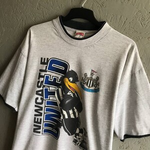 Puede incluir: Camiseta gris con un p&aacute;jaro de dibujos animados en blanco y negro que lleva una camiseta con las palabras "Newcastle United" en ella. La camiseta tiene un ribete negro alrededor del cuello y las mangas.
