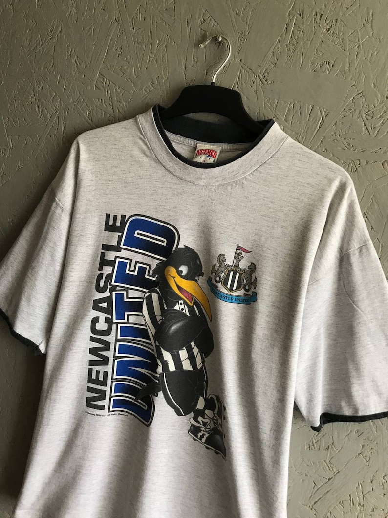 Puede incluir: Camiseta gris con un p&aacute;jaro de dibujos animados en blanco y negro que lleva una camiseta con las palabras "Newcastle United" en ella. La camiseta tiene un ribete negro alrededor del cuello y las mangas.