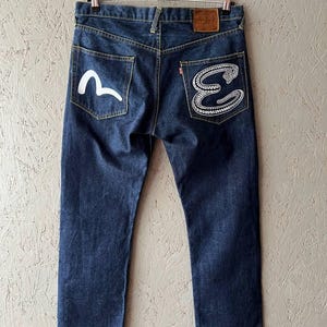 Può includere: Jeans in denim blu scuro con disegni ricamati bianchi sulle tasche posteriori. Una tasca presenta una "E" stilizzata e un disegno a serpente, l'altra un'onda bianca. Una toppa in pelle marrone è visibile in vita.