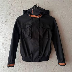 May include: Black jacket with a hood and two front pockets. The jacket has a brown leather strap detail on the hood and cuffs. The jacket is hanging on a white hanger against a light-colored wall.