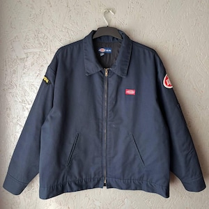 May include: A navy blue Dickies work jacket featuring a full-length front zipper, a pointed collar, and two slanted front pockets. The jacket includes a small red Dickies logo patch on the chest, a circular red and white patch on the left sleeve, and a black patch on the right sleeve.