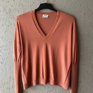 May include: A coral-colored V-neck sweater with long sleeves, hanging on a black hanger. The sweater has a tag that reads "Acne Studios". The garment is made of a soft material.
