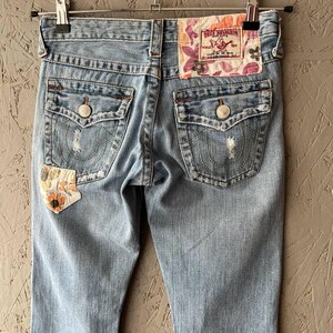 Puede incluir: Vaqueros de mezclilla azul claro con detalles desgastados, costuras decorativas y un parche floral. Los bolsillos traseros tienen cierres de bot&oacute;n. La etiqueta de la marca True Religion es visible en la cintura trasera.