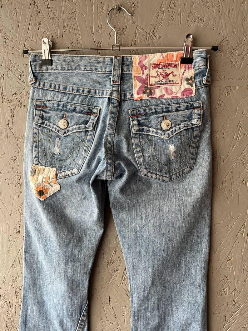 Puede incluir: Vaqueros de mezclilla azul claro con detalles desgastados, costuras decorativas y un parche floral. Los bolsillos traseros tienen cierres de bot&oacute;n. La etiqueta de la marca True Religion es visible en la cintura trasera.