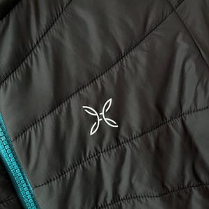 May include: Close-up of a black quilted jacket with a turquoise zip. The jacket features a white logo on the upper left chest. The stitching creates a diamond pattern across the jacket's surface.