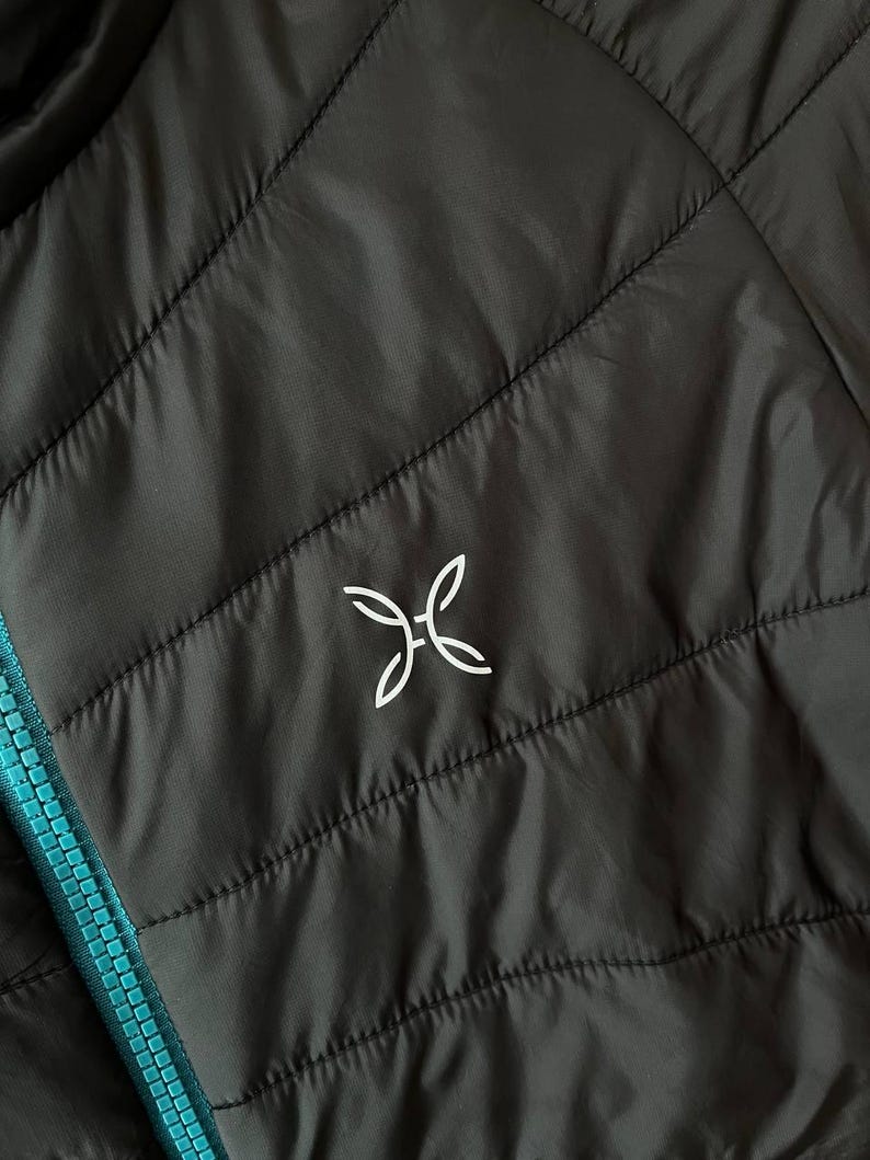 May include: Close-up of a black quilted jacket with a turquoise zip. The jacket features a white logo on the upper left chest. The stitching creates a diamond pattern across the jacket's surface.