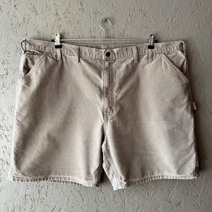 May include: Beige work shorts hanging on a metal hanger. The shorts feature multiple pockets, a button fastening, and a robust fabric. The shorts are a neutral colour, suitable for various outfits and activities.
