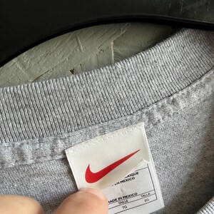 May include: A white tag with a red Nike swoosh logo and the text "Made in Mexico" in black. The tag also has the words "Taille" and "Talla" in black, with "TG" and "EG" underneath.