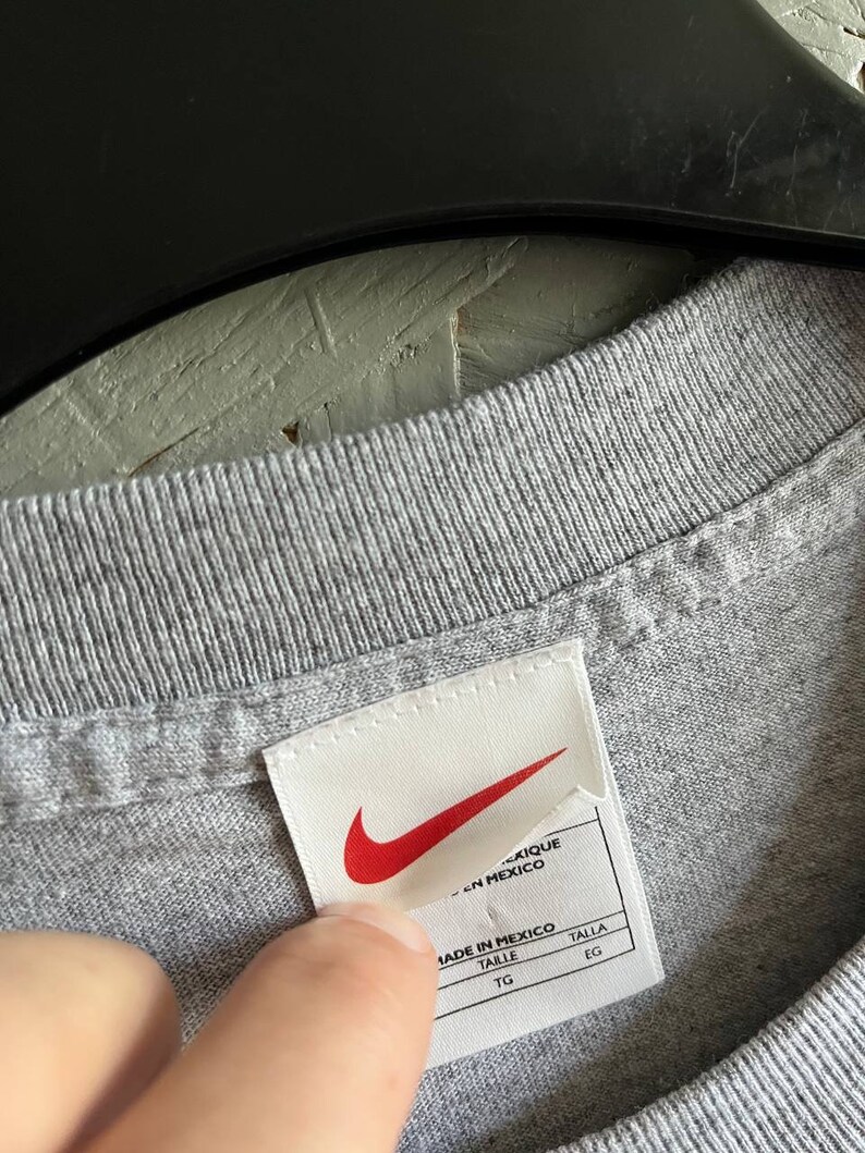 May include: A white tag with a red Nike swoosh logo and the text "Made in Mexico" in black. The tag also has the words "Taille" and "Talla" in black, with "TG" and "EG" underneath.