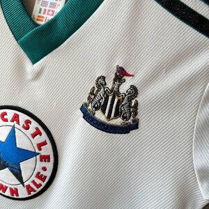 K&ouml;nnte beinhalten: Wei&szlig;es Fu&szlig;balltrikot mit gr&uuml;nem V-Ausschnitt und schwarzen Streifen auf der Schulter. Mit dem Newcastle United-Wappen und einem "Town Ale"-Patch. Ein Adidas-Etikett ist sichtbar.