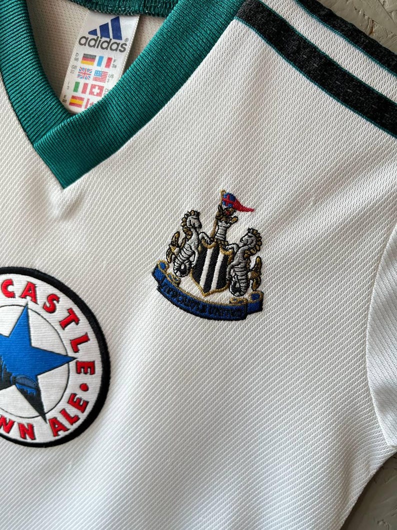 K&ouml;nnte beinhalten: Wei&szlig;es Fu&szlig;balltrikot mit gr&uuml;nem V-Ausschnitt und schwarzen Streifen auf der Schulter. Mit dem Newcastle United-Wappen und einem "Town Ale"-Patch. Ein Adidas-Etikett ist sichtbar.