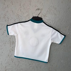 K&ouml;nnte beinhalten: Wei&szlig;es Sportshirt mit t&uuml;rkisfarbenen und schwarzen Zierleisten. Das kurz&auml;rmelige Shirt hat einen t&uuml;rkisfarbenen Kragen und eine Zierleiste um die &Auml;rmel. Schwarze Streifen verlaufen &uuml;ber die Schultern. Das Shirt h&auml;ngt an einem schwarzen Kleiderb&uuml;gel vor grauem Hintergrund.