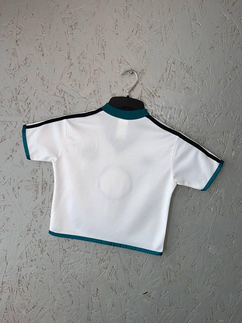 K&ouml;nnte beinhalten: Wei&szlig;es Sportshirt mit t&uuml;rkisfarbenen und schwarzen Zierleisten. Das kurz&auml;rmelige Shirt hat einen t&uuml;rkisfarbenen Kragen und eine Zierleiste um die &Auml;rmel. Schwarze Streifen verlaufen &uuml;ber die Schultern. Das Shirt h&auml;ngt an einem schwarzen Kleiderb&uuml;gel vor grauem Hintergrund.