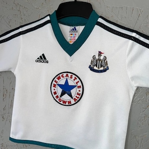 K&ouml;nnte beinhalten: Wei&szlig;es Fu&szlig;balltrikot mit schwarzen Streifen auf den Schultern und einem gr&uuml;nen Kragen. Mit Adidas-Logo, Vereinswappen und einem runden Aufn&auml;her mit Text und blauem Stern. Das Trikot h&auml;ngt an einem schwarzen Kleiderb&uuml;gel.