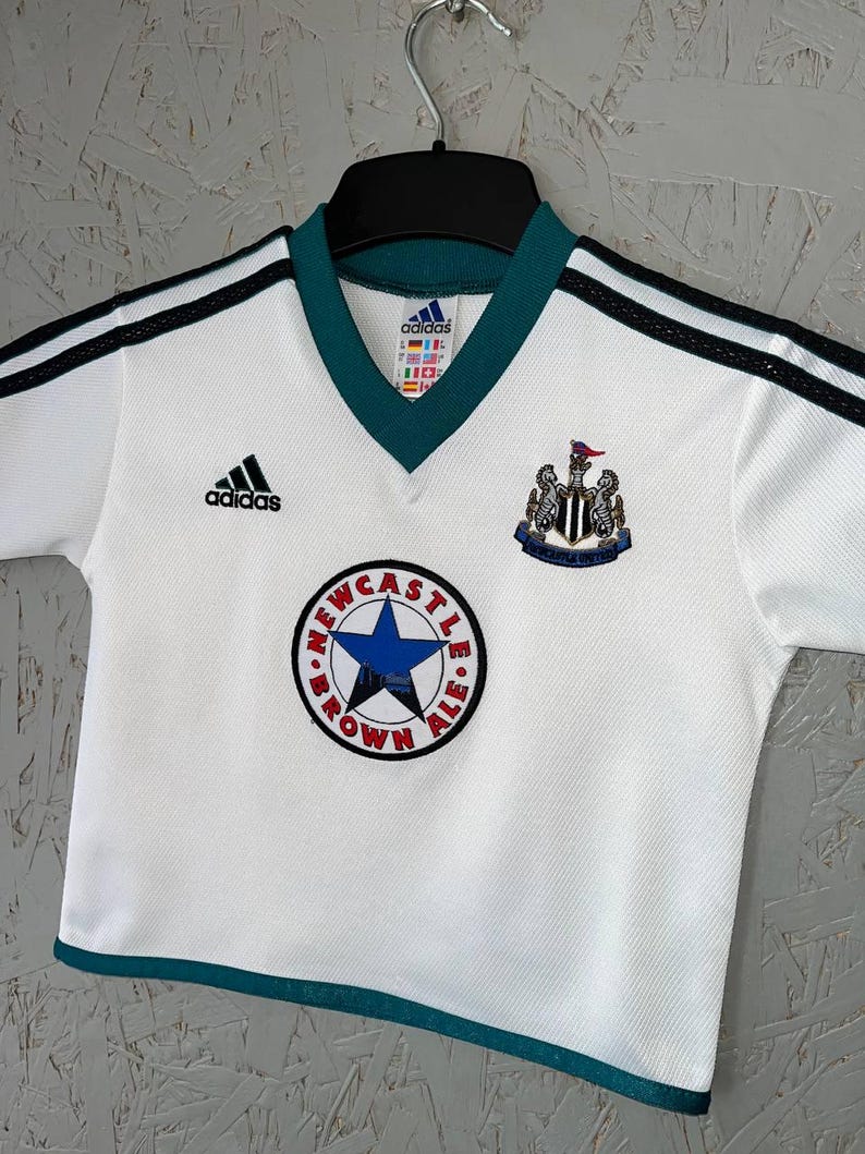 K&ouml;nnte beinhalten: Wei&szlig;es Fu&szlig;balltrikot mit schwarzen Streifen auf den Schultern und einem gr&uuml;nen Kragen. Mit Adidas-Logo, Vereinswappen und einem runden Aufn&auml;her mit Text und blauem Stern. Das Trikot h&auml;ngt an einem schwarzen Kleiderb&uuml;gel.