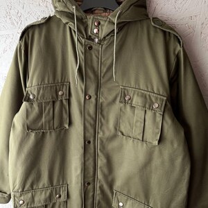 K&ouml;nnte beinhalten: Eine olivgr&uuml;ne Kapuzenjacke mit Knopfleiste auf der Vorderseite, vier gro&szlig;en Pattentaschen mit Metalldruckkn&ouml;pfen, Schulterklappen und einem gesteppten braunen Innenfutter. Die Jacke verf&uuml;gt &uuml;ber eine Kapuze mit Kordelzug und lange &Auml;rmel.