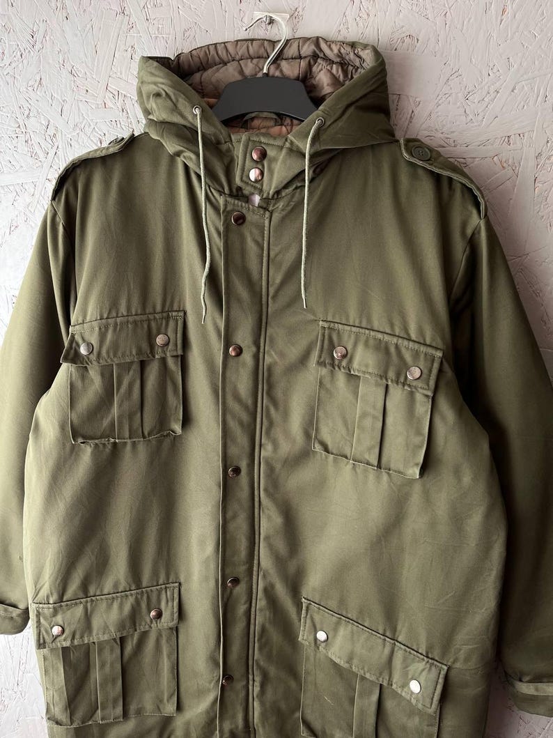 K&ouml;nnte beinhalten: Eine olivgr&uuml;ne Kapuzenjacke mit Knopfleiste auf der Vorderseite, vier gro&szlig;en Pattentaschen mit Metalldruckkn&ouml;pfen, Schulterklappen und einem gesteppten braunen Innenfutter. Die Jacke verf&uuml;gt &uuml;ber eine Kapuze mit Kordelzug und lange &Auml;rmel.