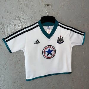 K&ouml;nnte beinhalten: Wei&szlig;es Fu&szlig;balltrikot mit schwarzen Streifen auf den &Auml;rmeln und t&uuml;rkisfarbenen Verzierungen. Das Trikot zeigt das Adidas-Logo, ein Newcastle Brown Ale-Logo und ein Teamwappen. Das Trikot h&auml;ngt an einem schwarzen Kleiderb&uuml;gel.
