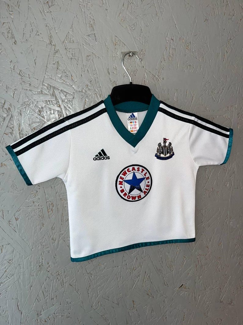 K&ouml;nnte beinhalten: Wei&szlig;es Fu&szlig;balltrikot mit schwarzen Streifen auf den &Auml;rmeln und t&uuml;rkisfarbenen Verzierungen. Das Trikot zeigt das Adidas-Logo, ein Newcastle Brown Ale-Logo und ein Teamwappen. Das Trikot h&auml;ngt an einem schwarzen Kleiderb&uuml;gel.
