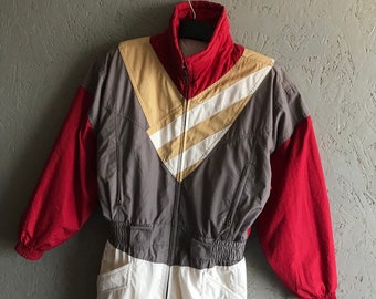 Könnte beinhalten: Ein Vintage-Ski-Overall in Rot, Grau, Weiß und Beige mit Reißverschluss. Der Overall hat ein geometrisches Muster und einen hohen Kragen.
