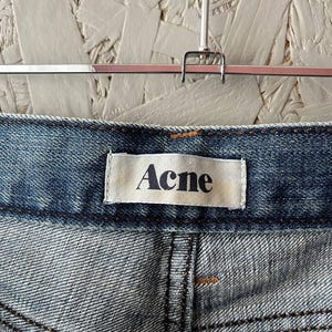 以下が含まれることがあります： 白いラベルに「Acne」のブランド名が入った、青いデニムジーンズのクローズアップ。 ジーンズは、明るい色のテクスチャード加工された背景に対して、シルバーのハンガーに吊るされています。 ステッチが見えます。