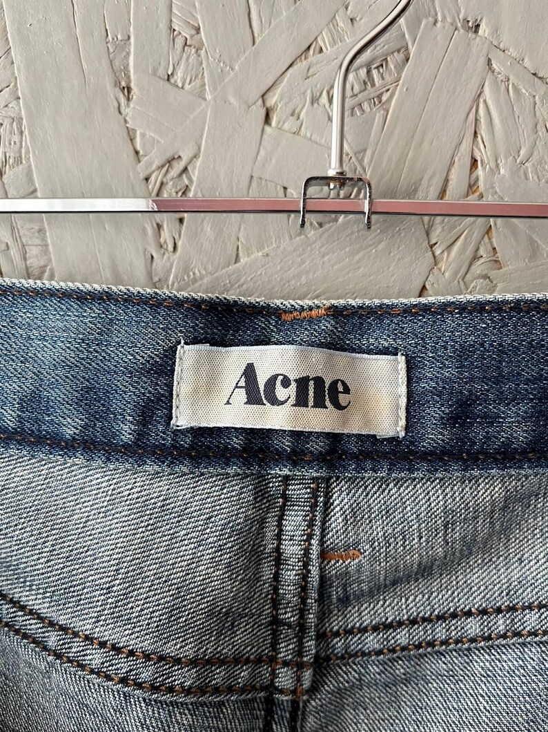 以下が含まれることがあります： 白いラベルに「Acne」のブランド名が入った、青いデニムジーンズのクローズアップ。 ジーンズは、明るい色のテクスチャード加工された背景に対して、シルバーのハンガーに吊るされています。 ステッチが見えます。