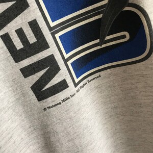Puede incluir: Camiseta gris con un gr&aacute;fico azul y negro que dice "AVEN." El gr&aacute;fico tiene un contorno blanco. El texto "Nutmeg Mills Inc. All Rights Reserved" est&aacute; impreso debajo del gr&aacute;fico.