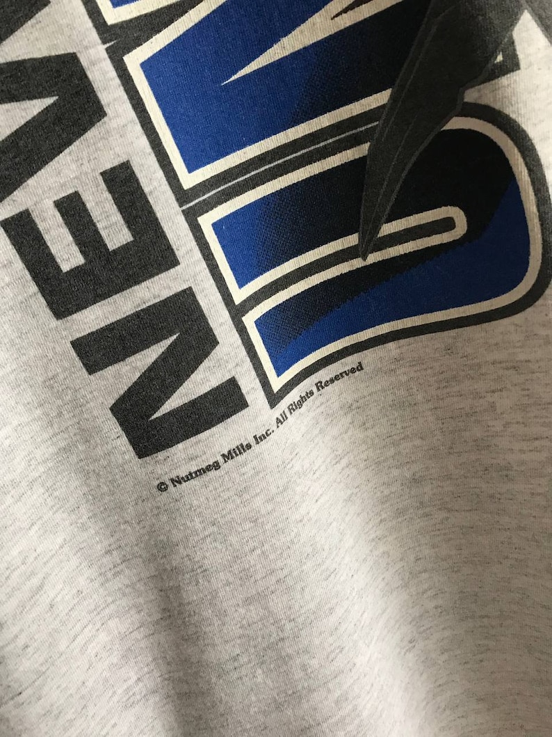 Puede incluir: Camiseta gris con un gr&aacute;fico azul y negro que dice "AVEN." El gr&aacute;fico tiene un contorno blanco. El texto "Nutmeg Mills Inc. All Rights Reserved" est&aacute; impreso debajo del gr&aacute;fico.