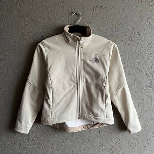 Può includere: Una giacca in softshell beige chiaro con chiusura a zip e un logo The North Face sul petto sinistro.