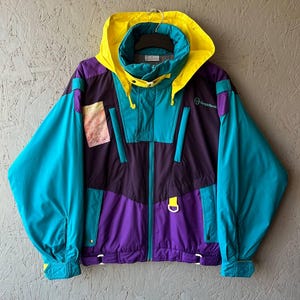 Op de afbeelding: Een vintage jas met kleurblokken en een capuchon. De jas heeft een levendig kleurenschema van teal, paars, geel en marineblauw. Het heeft een ritssluiting, een hoge kraag en een kleine patch aan de linkerkant. Retro stijl.