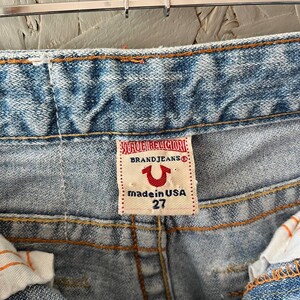 Puede incluir: Primer plano de vaqueros de mezclilla azul claro con costuras naranjas. La etiqueta de la marca True Religion es visible, con el texto "BRAND JEANS", un logotipo de herradura y "made in USA 27". Los vaqueros est&aacute;n colgados de una percha met&aacute;lica.