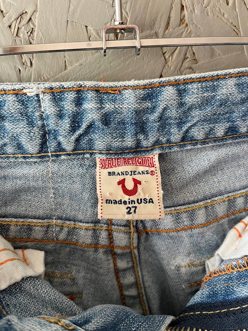 Puede incluir: Primer plano de vaqueros de mezclilla azul claro con costuras naranjas. La etiqueta de la marca True Religion es visible, con el texto "BRAND JEANS", un logotipo de herradura y "made in USA 27". Los vaqueros est&aacute;n colgados de una percha met&aacute;lica.
