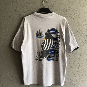 Puede incluir: Una camiseta gris con un dibujo animado en blanco y negro de un ping&uuml;ino patinando con un uniforme de &aacute;rbitro. El texto "NEWCASTLE UNITED" est&aacute; impreso verticalmente en la camiseta.